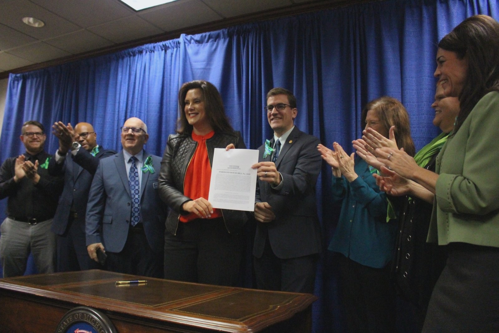 Governor Gretchen Whitmer holds up an enrolled Michigan House bill after signing it, flanked by legislators and counseling advocates applauding