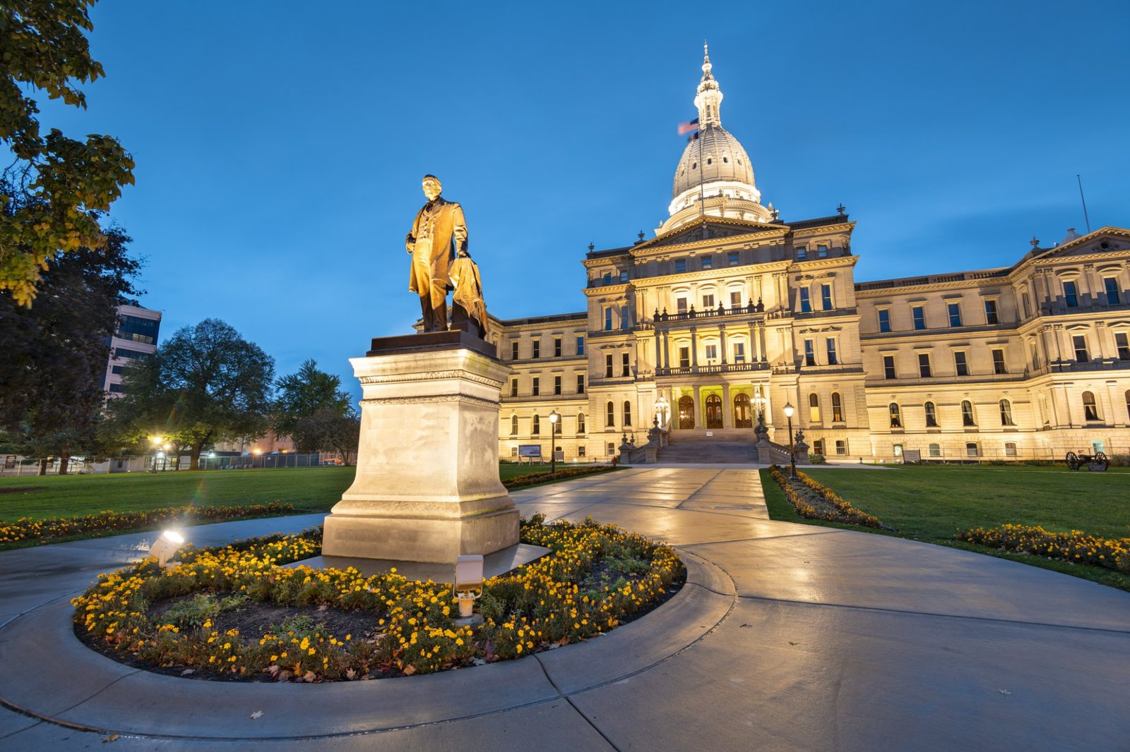 The Michigan State Capitol in Lansing at dusk, lit from below, with the Austin Blair statue and marigold beds in the foreground