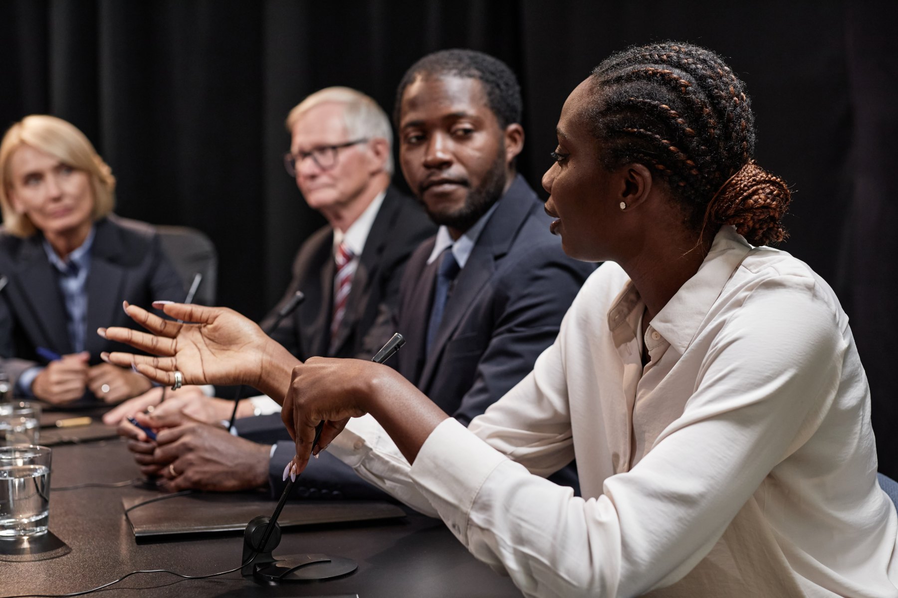 A witness speaking into a table microphone at a legislative hearing, gesturing to make a point, with three committee members listening