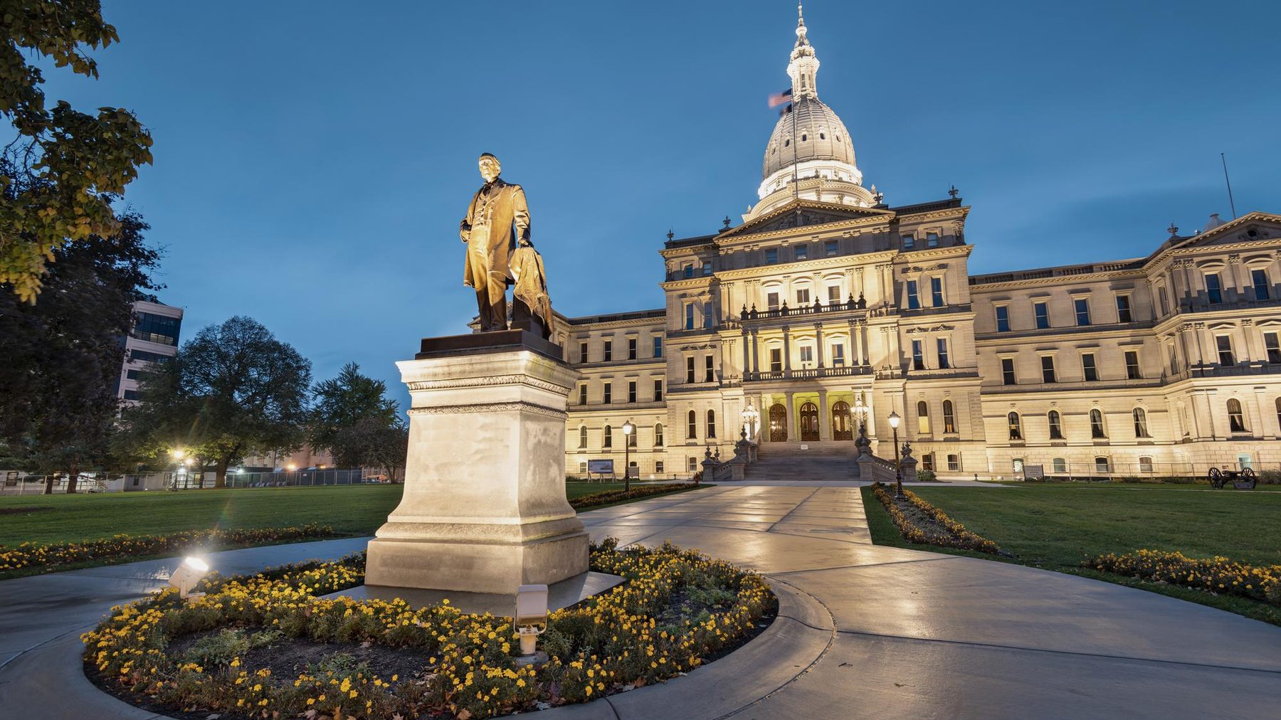 The Michigan State Capitol lit at dusk, with the Austin Blair statue and flowerbeds in the foreground