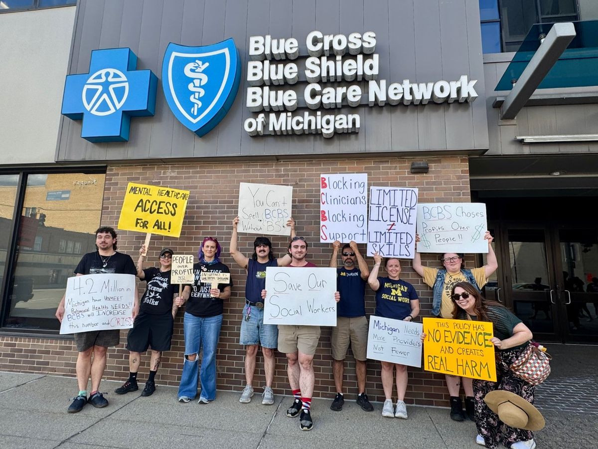 Eleven clinicians and supporters lined up beneath the Blue Cross Blue Shield Blue Care Network of Michigan sign, holding hand-lettered signs reading Mental Healthcare Access For All, Limited License does not equal Limited Skills, Save Our Social Workers, Michigan Needs More Providers Not Fewer, and This Policy Has No Evidence And Creates Real Harm