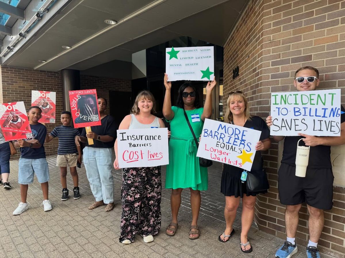 A counselor standing alone outside a Blue Care Network of Michigan building holding a hand-lettered sign reading Keep Care Accessible