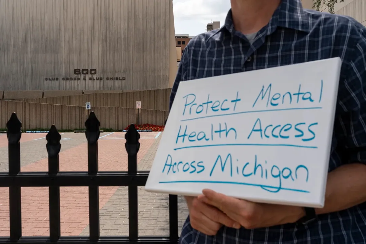 A person holds a whiteboard sign reading "Protect Mental Health Access Across Michigan" in front of a building marked 600 Blue Cross Blue Shield.