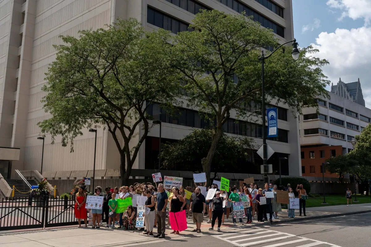 A wide view of about forty people with signs lined along the sidewalk and fence outside a large office building at a street corner.