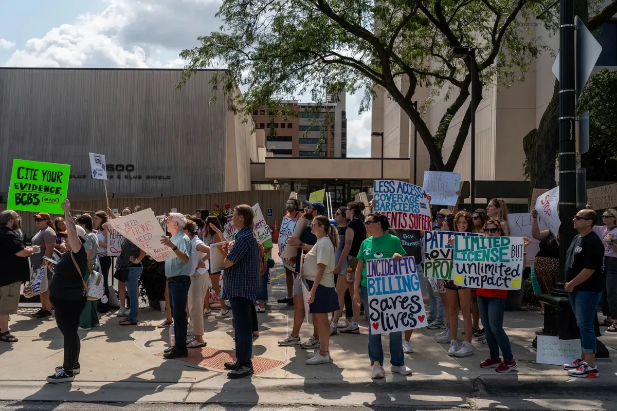 Demonstrators with signs fill the sidewalk in front of the 600 Blue Cross Blue Shield building entrance; one sign reads "CITE YOUR EVIDENCE, BCBS."