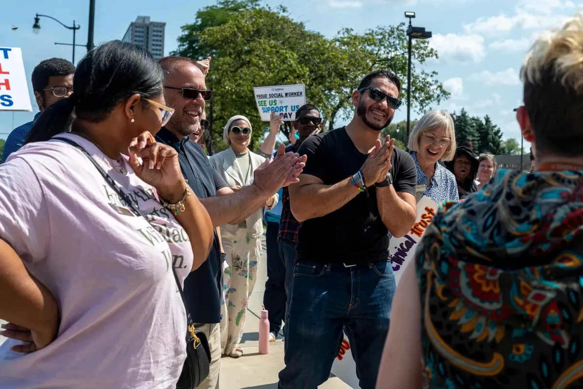 People in a circle applaud during the rally; a sign behind them reads "PROUD SOCIAL WORKER, SEE US, SUPPORT US, PAY US."