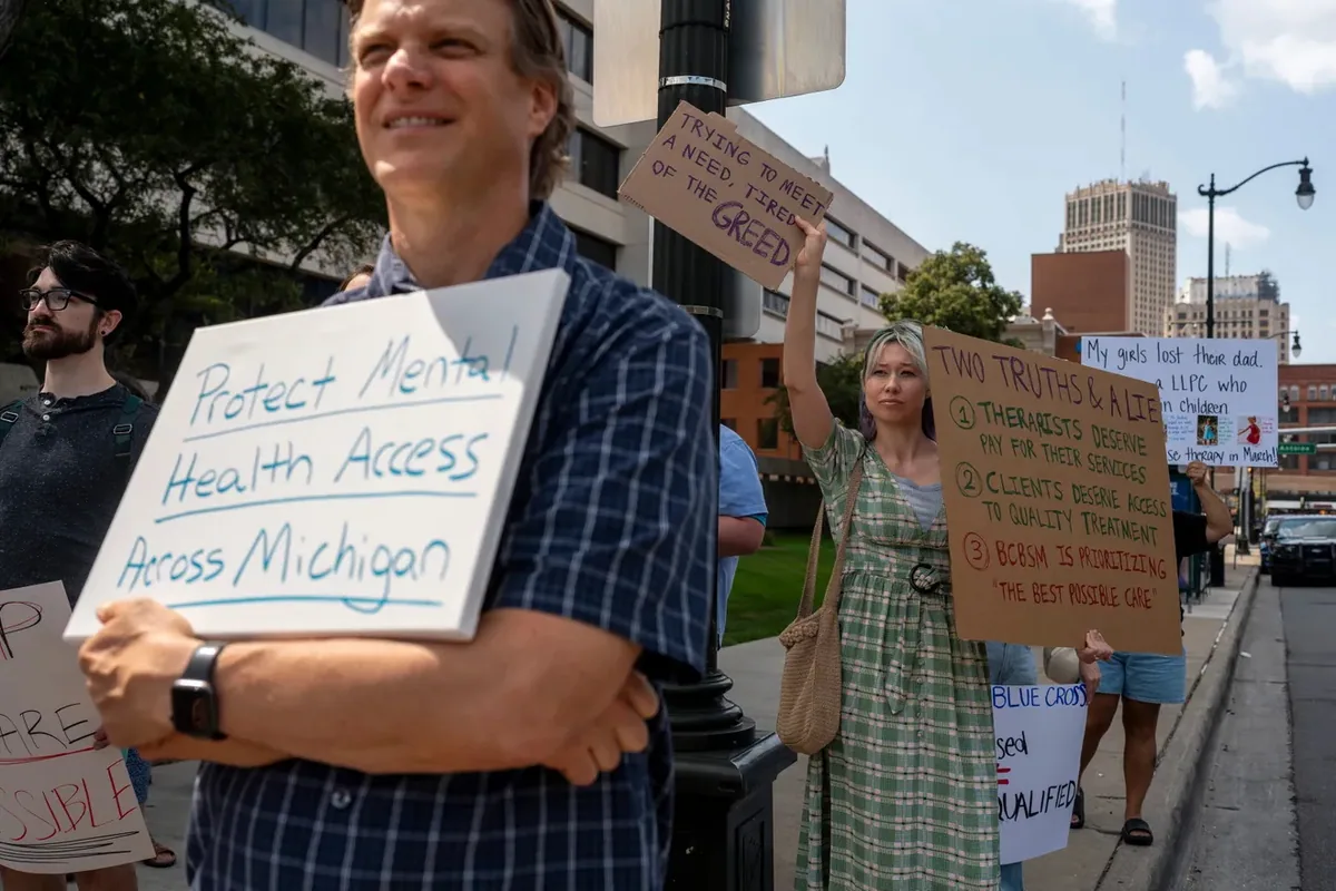 A person holds a whiteboard reading "Protect Mental Health Access Across Michigan" while others behind hold signs including "TRYING TO MEET A NEED, TIRED OF THE GREED."
