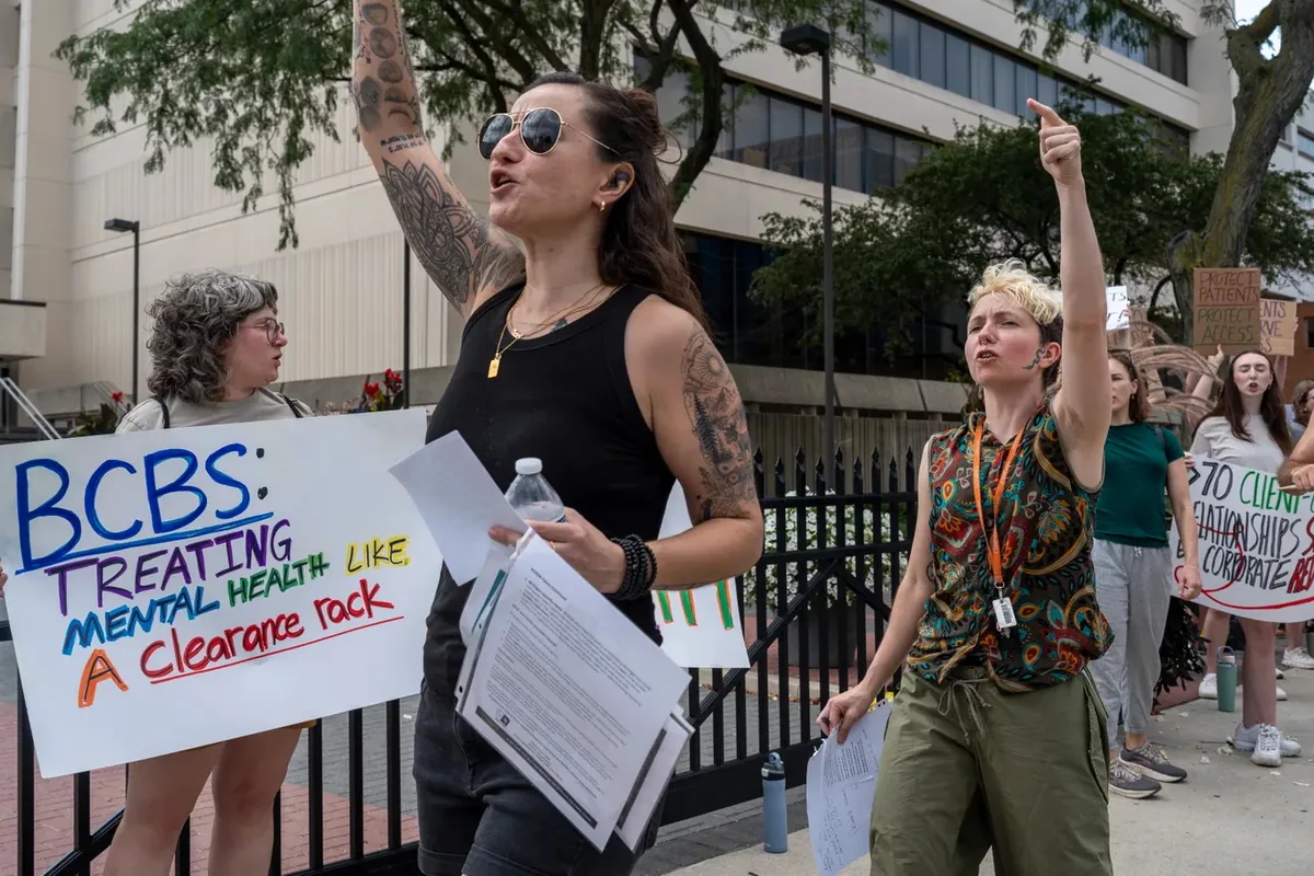 Two people chant with raised arms and papers in hand beside a sign reading "BCBS: treating mental health like a clearance rack."