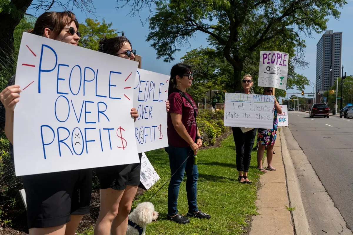 Demonstrators line a grass strip beside a roadway holding matching "PEOPLE OVER PROFITS" signs and one reading "CHOICE MATTERS, Let Clients Make the Choice."