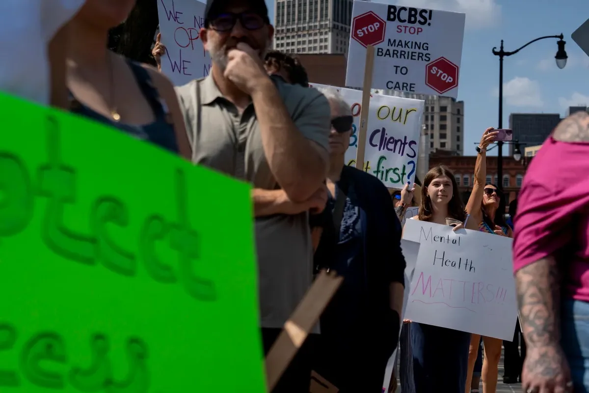 Demonstrators hold signs reading "BCBS, stop making barriers to care" and "Mental Health Matters" at the roadside.