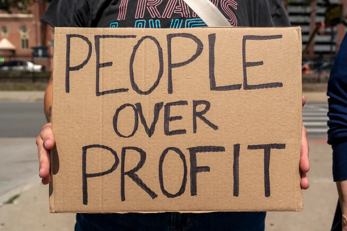 A close view of two hands holding a cardboard sign lettered "PEOPLE OVER PROFIT" at chest height on a street.