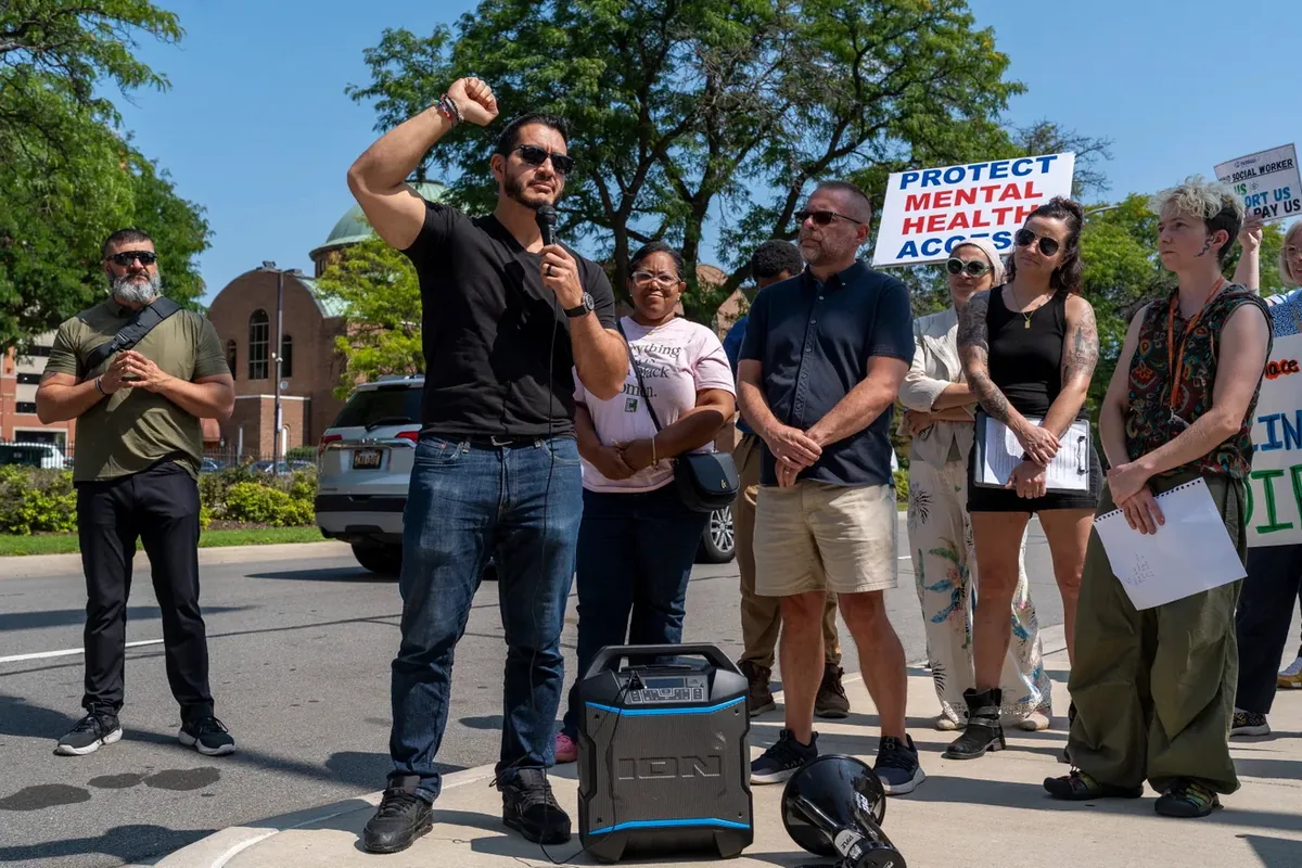 A speaker with a microphone raises a fist while addressing the rally, with a portable speaker and bullhorn on the pavement and supporters standing behind.
