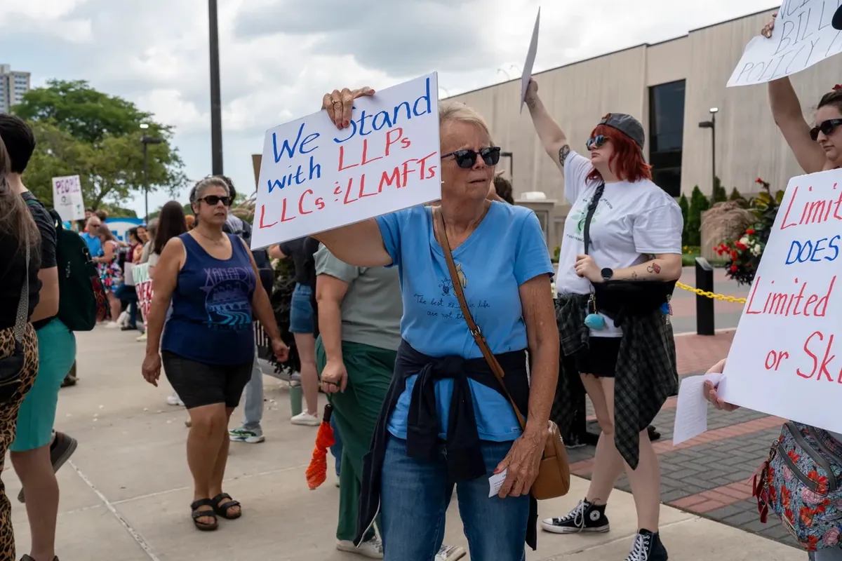 A person holds a sign reading "We Stand with LLPCs, LLMSWs and LLMFTs" while others walk and raise papers on the sidewalk behind.