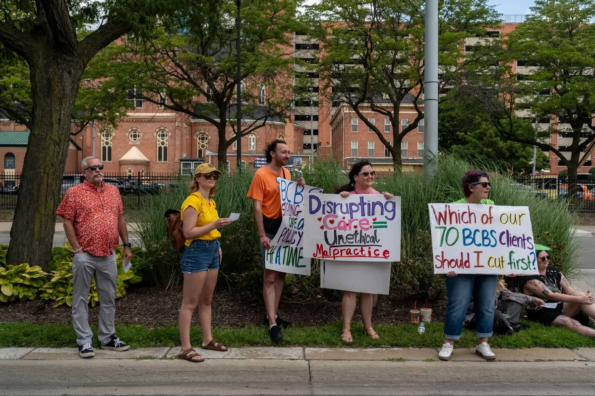 Five demonstrators stand along a landscaped roadside with signs including "Disrupting Care = Unethical Malpractice" and "Which of our 70 BCBS clients should I cut first?"