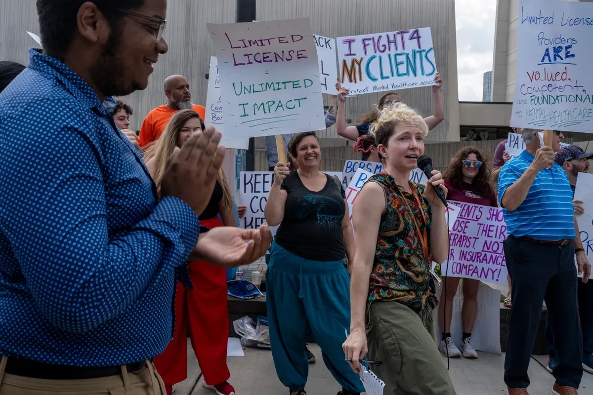 A person speaks into a microphone while others clap and hold signs reading "I FIGHT 4 MY CLIENTS" and "LIMITED LICENSE = UNLIMITED IMPACT."