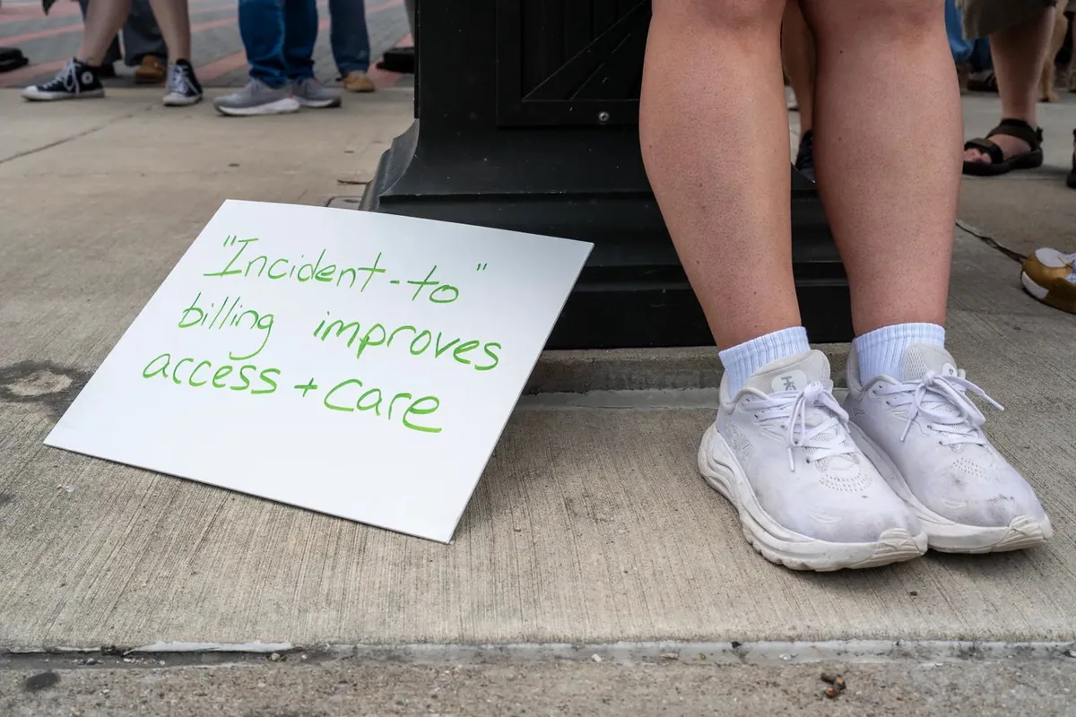 A sign reading "Incident-to billing improves access and care" leans against a lamppost base beside a person seated on a step.