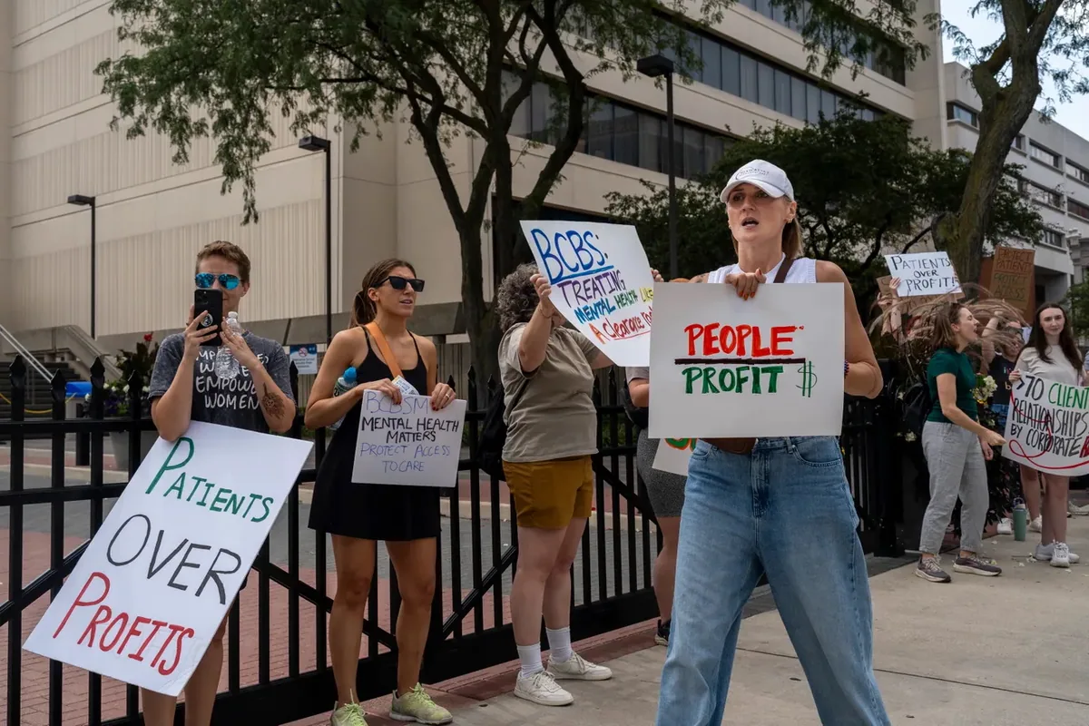 Demonstrators along an iron fence hold signs reading "PEOPLE OVER PROFIT" and "Patients OVER Profits" as one records on a phone.