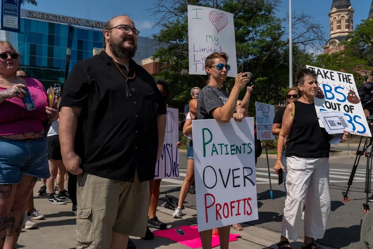 Demonstrators stand at a crosswalk holding signs reading "Patients OVER Profits," "TAKE THE BS OUT OF BCBS" and "I love my Limited License Therapist."