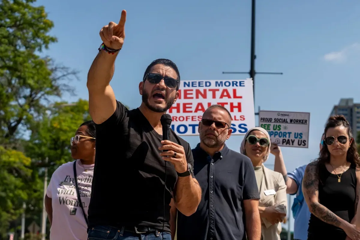 A speaker holding a microphone points upward while addressing the crowd, with signs behind reading "WE NEED MORE MENTAL HEALTH PROFESSIONALS NOT LESS."