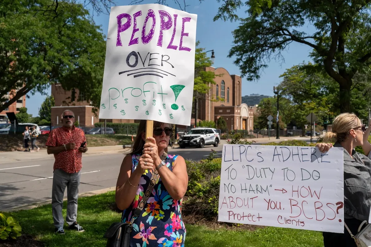A person holds a "PEOPLE OVER profit" sign on a wooden stake beside another reading "LLPCs adhere to duty to do no harm. How about you, BCBS?"