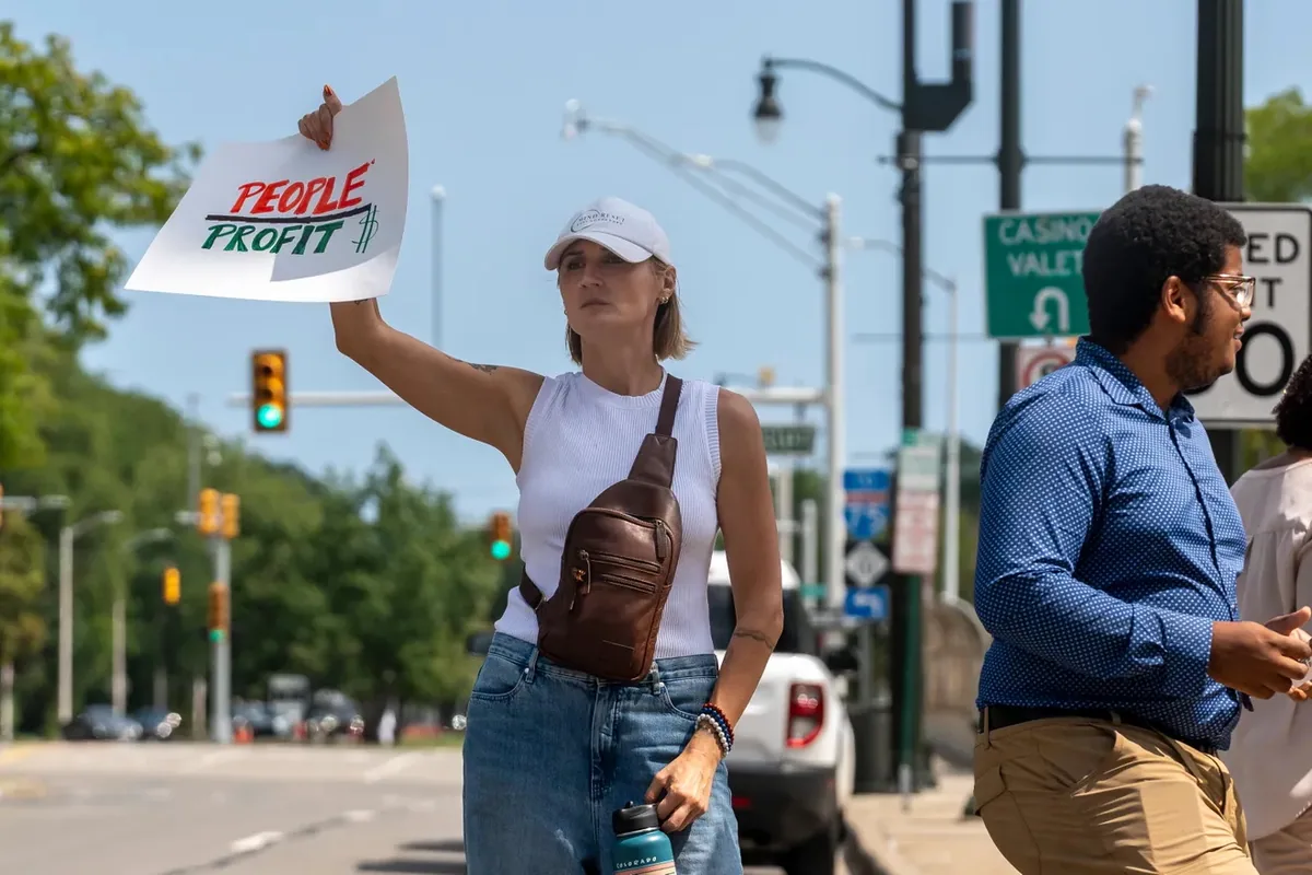 A person raises a paper sign reading "PEOPLE OVER PROFIT" toward passing traffic on a downtown street.
