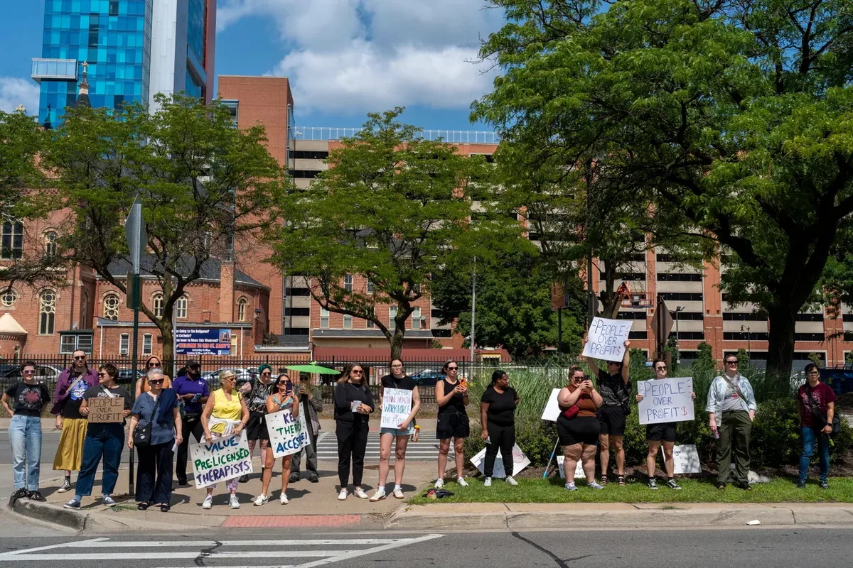A wide shot of roughly twenty demonstrators spread along a curb and median with signs including "PEOPLE OVER PROFITS" and "VALUE PRE-LICENSED THERAPISTS."