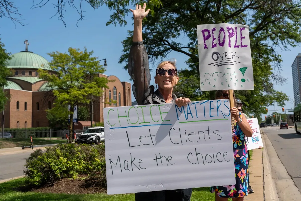 A person raises one arm while holding a sign reading "CHOICE MATTERS, Let Clients Make the Choice," with a "PEOPLE OVER profit" sign raised nearby.