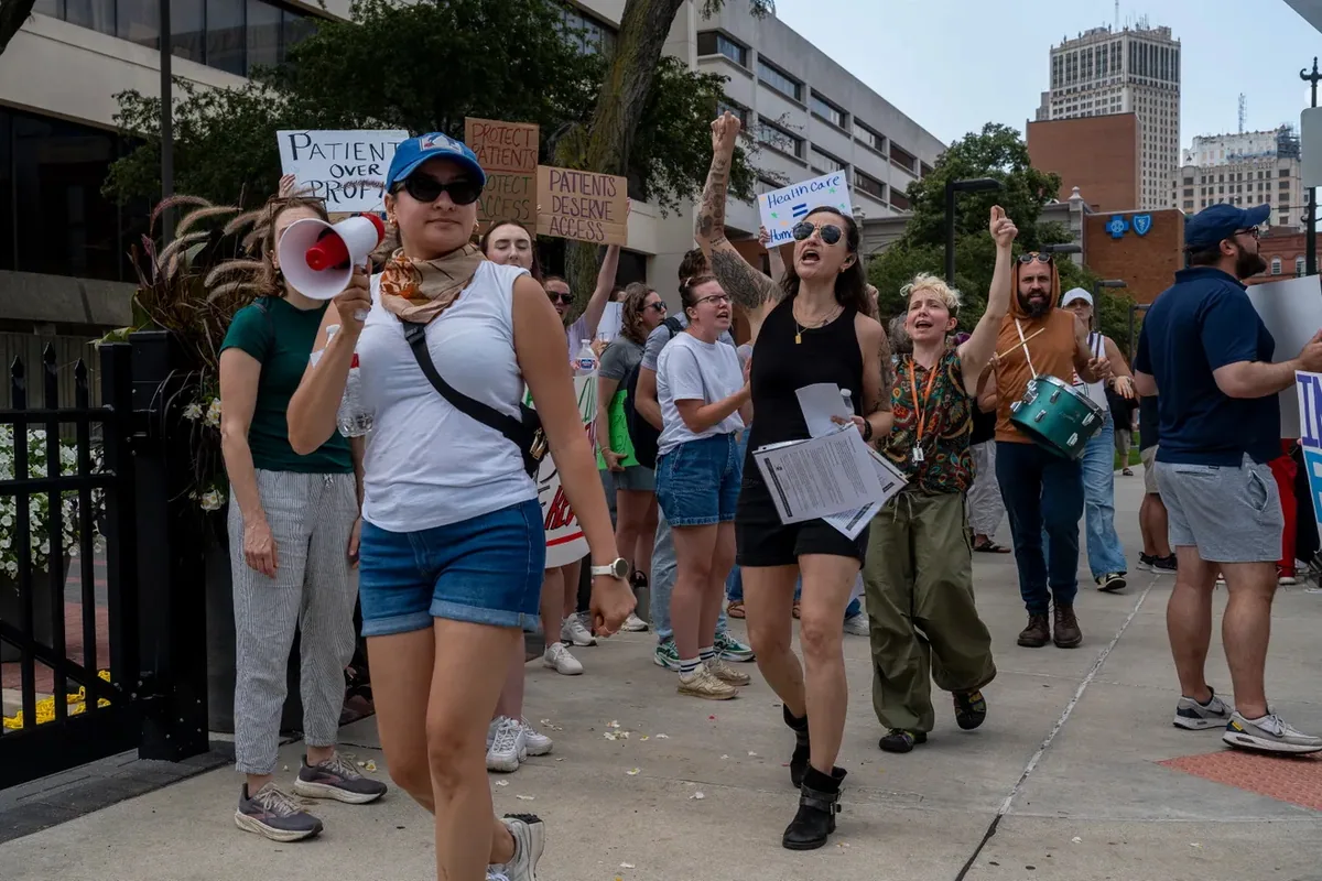 Marchers move along the sidewalk chanting, one using a bullhorn and another playing a drum, with signs reading "PATIENTS DESERVE ACCESS" and "PROTECT PATIENTS PROTECT ACCESS."