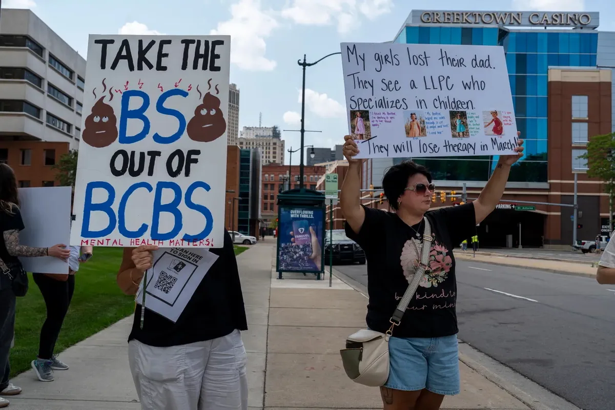 Two people hold signs on a sidewalk, one reading "TAKE THE BS OUT OF BCBS, MENTAL HEALTH MATTERS" and one describing a family who will lose their therapist in March.
