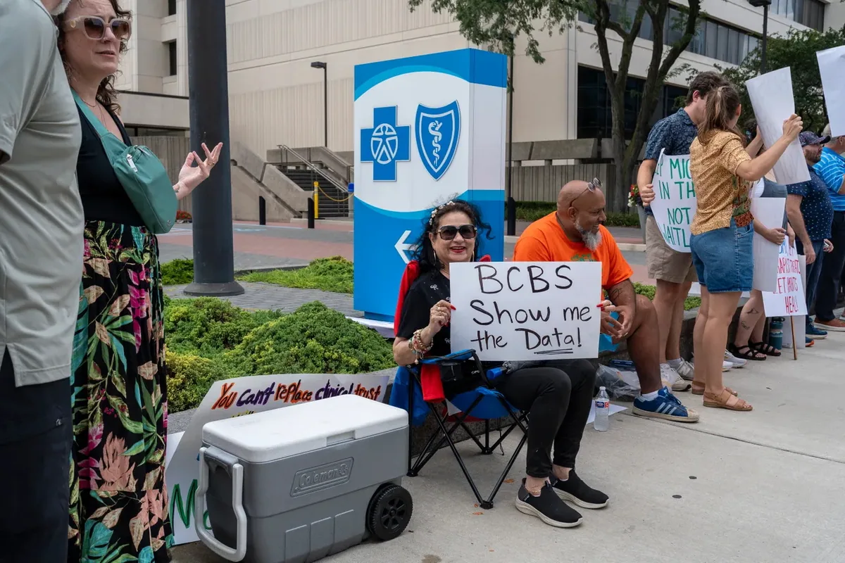 A person seated in a folding chair holds a sign reading "BCBS Show me the Data" beside a large Blue Cross Blue Shield logo sign.