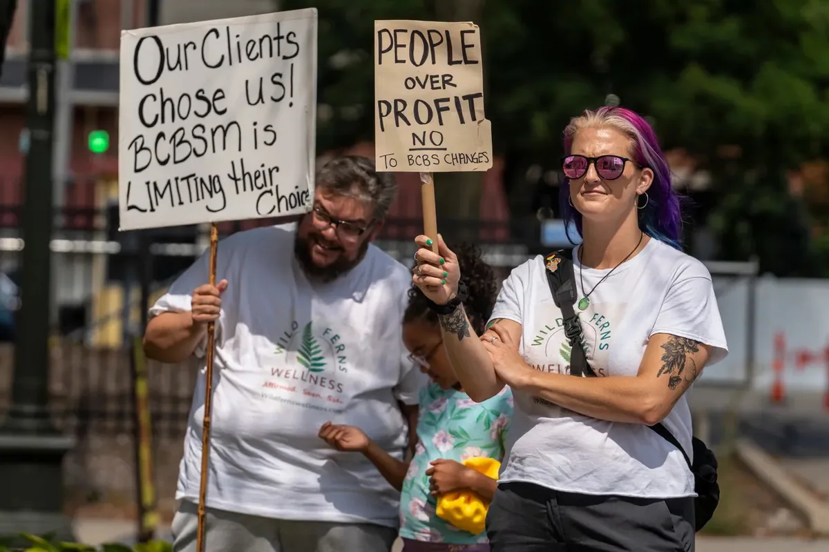 Two adults in matching Wild Ferns Wellness shirts stand with a child, holding signs reading "Our Clients Chose Us, BCBSM is limiting their choice."
