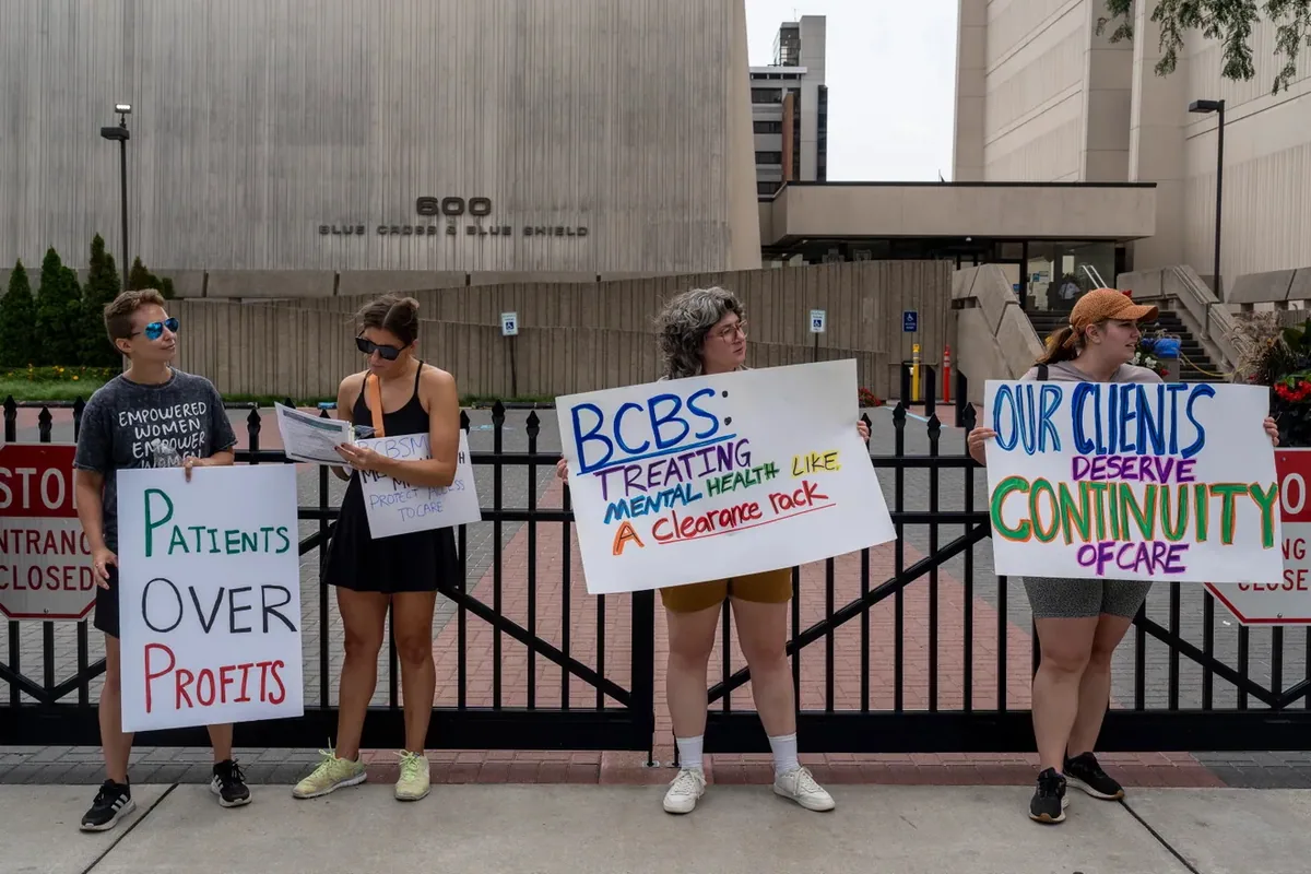 Four people hold signs along a fence in front of the 600 Blue Cross Blue Shield building, including "OUR CLIENTS DESERVE CONTINUITY OF CARE" and "Patients OVER Profits."