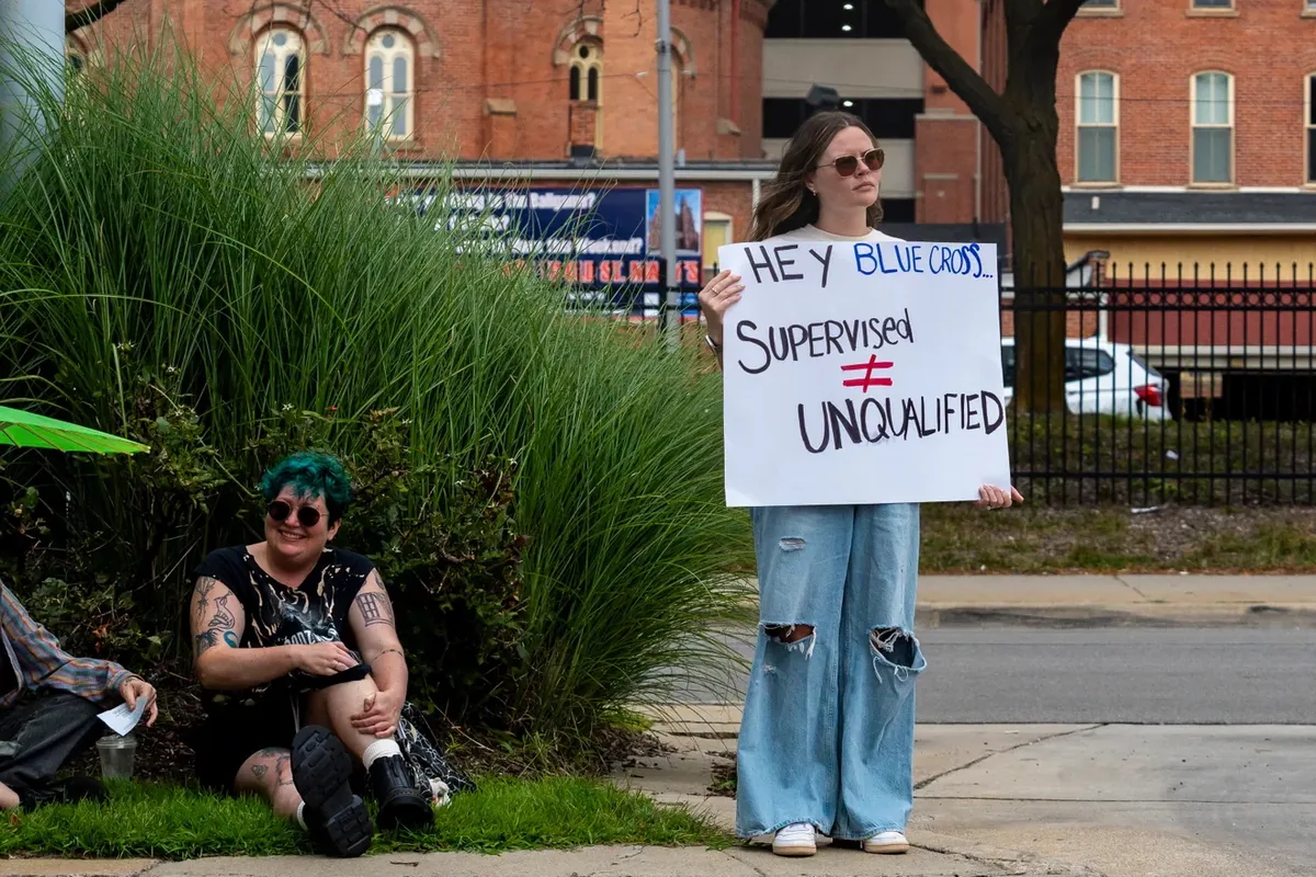 A person stands at the roadside holding a sign reading "HEY BLUE CROSS, SUPERVISED DOES NOT MEAN UNQUALIFIED."