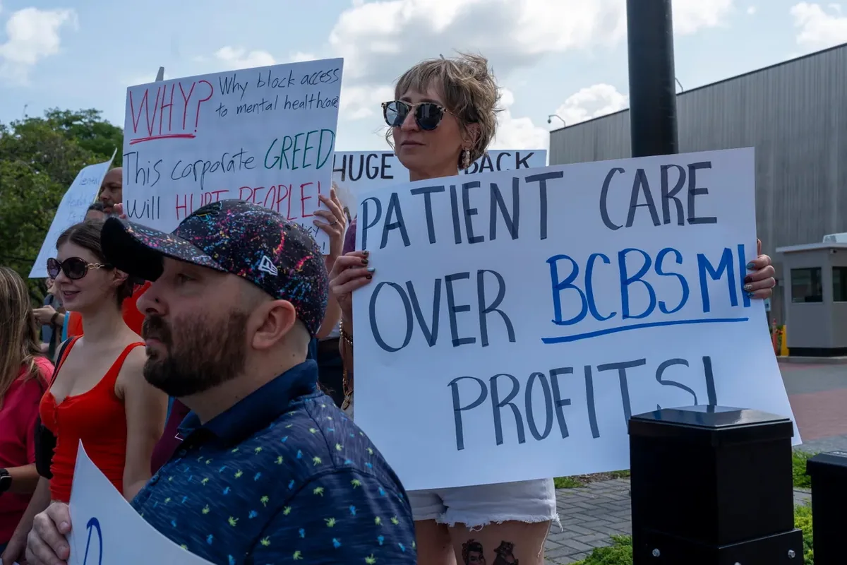 A person holds a sign reading "PATIENT CARE OVER BCBSMI PROFITS" beside another asking why access to mental health care is being blocked.