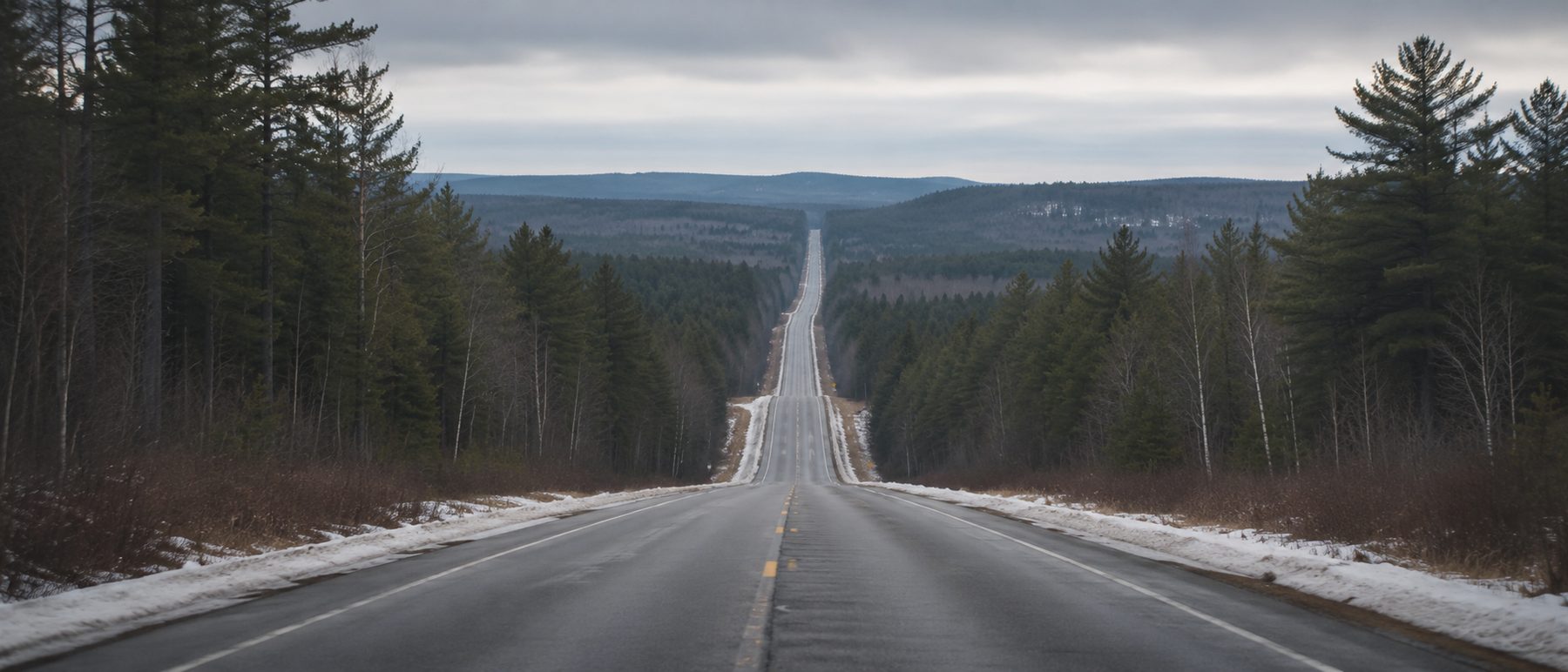 A two-lane highway receding through forest with patchy snow and no vehicles in sight